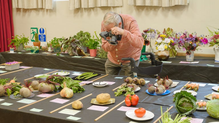 Photographer Martin Parr takes a photo at the Lacock Flower Show. He is bending over a table covered in produce, pointing his lens at a prize winning potato.